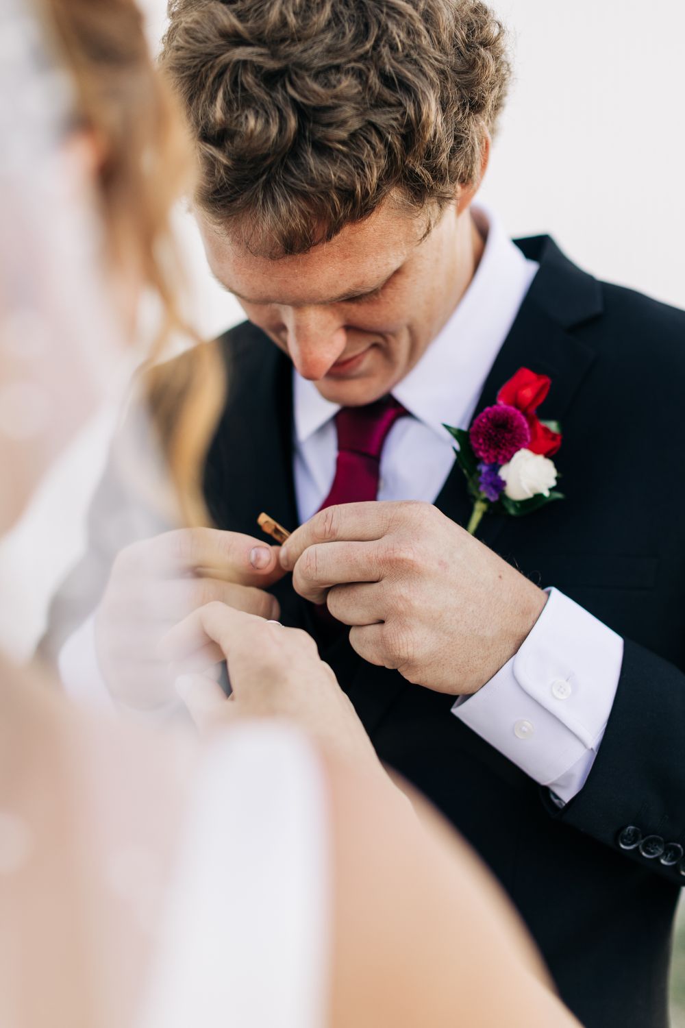 Groom in black suit examines wedding ring with boutonniere of red and purple flowers.