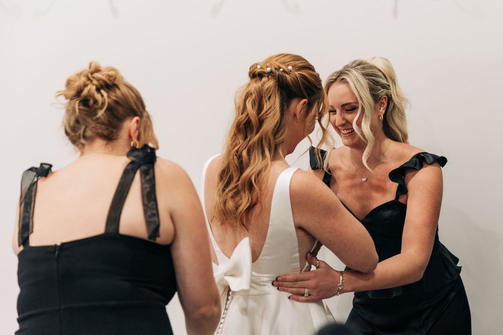 Bride in white gown with bridesmaids in black dresses sharing a joyful moment during wedding preparations.