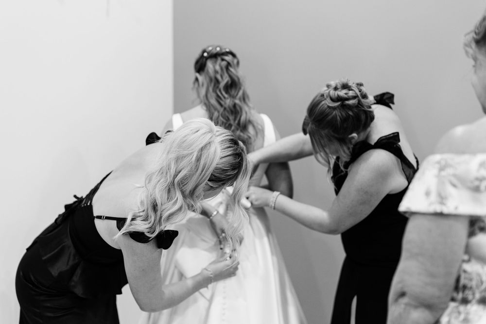 Bridesmaids in black dresses help a bride with her white wedding gown in a black and white photo.