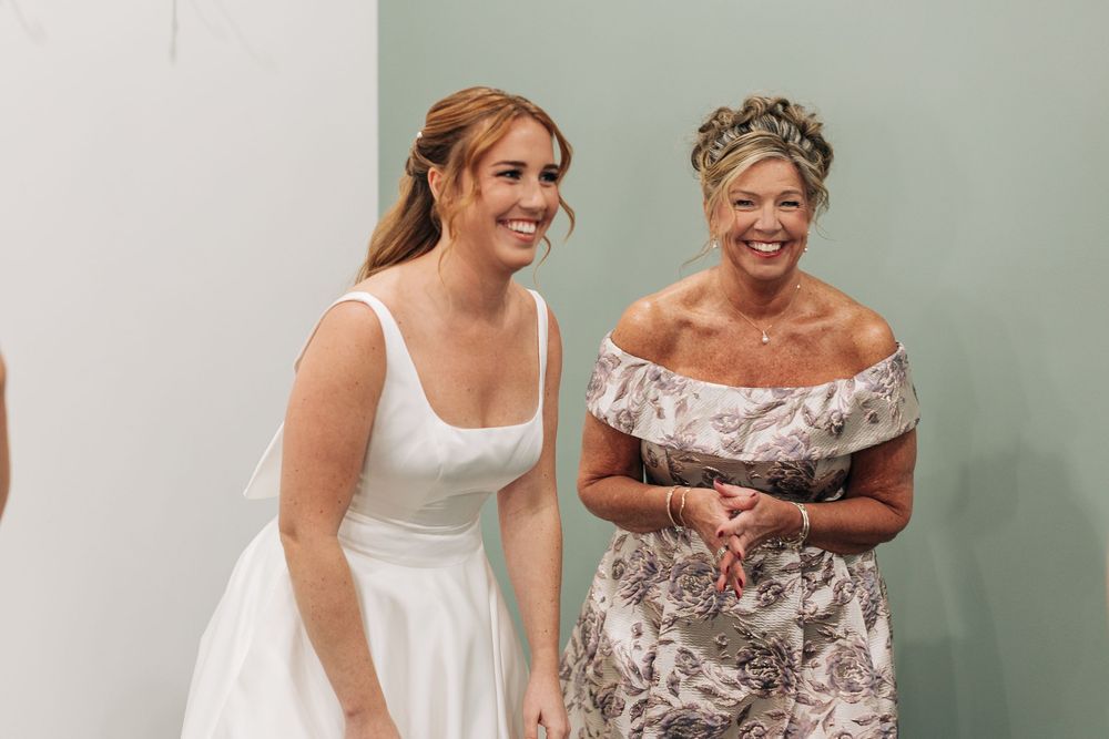 Bride in white dress laughing with older woman in floral off-shoulder dress at a wedding celebration.