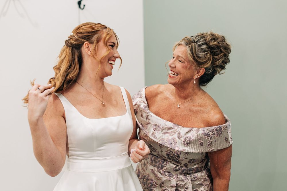 Bride in white gown smiling with older woman in floral off-shoulder dress at wedding celebration.