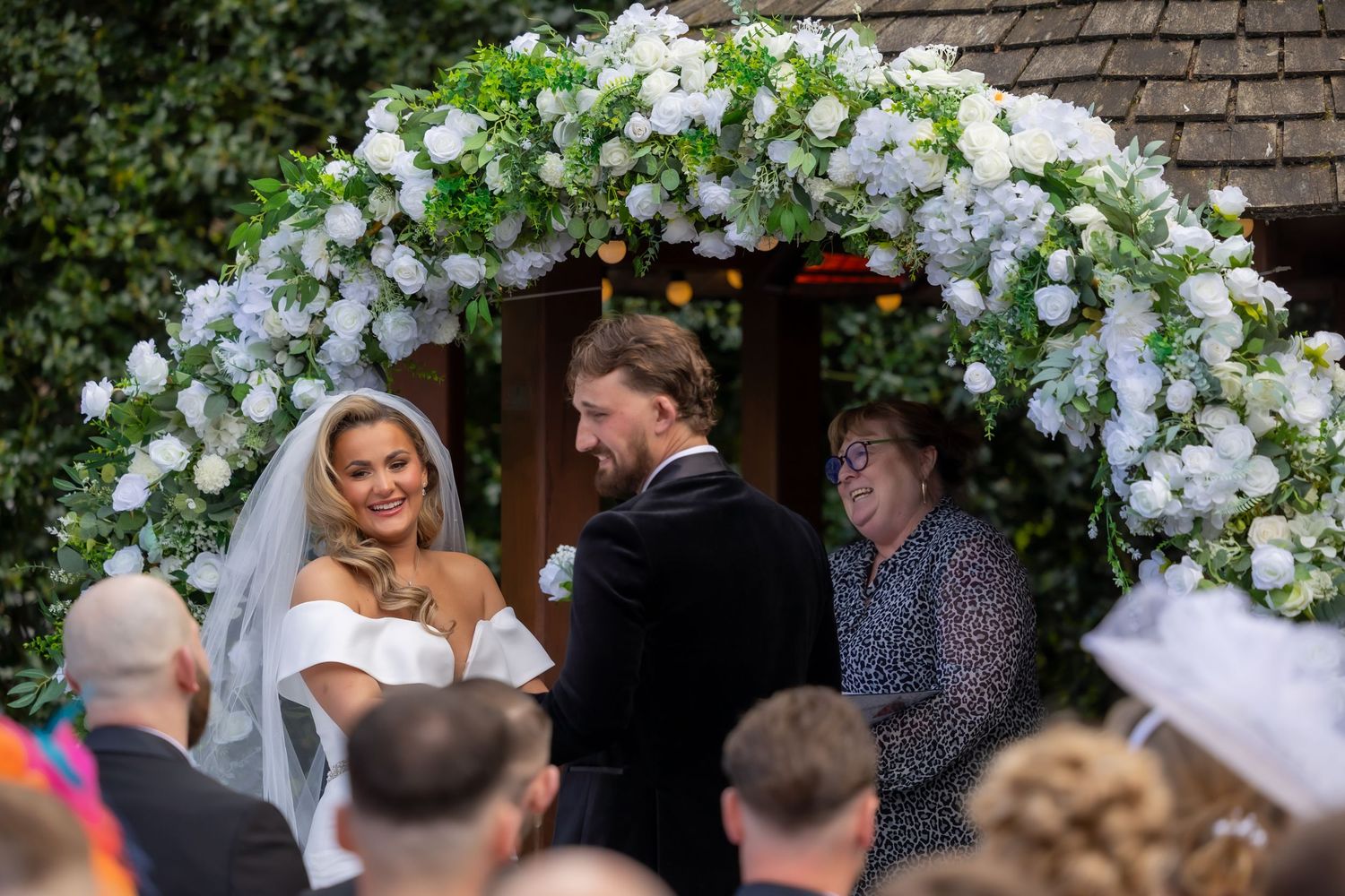 Bride and groom exchange vows under a floral arch  with white flowers at an outdoor wedding ceremony. Casual Captures Bannatynes Hotel.