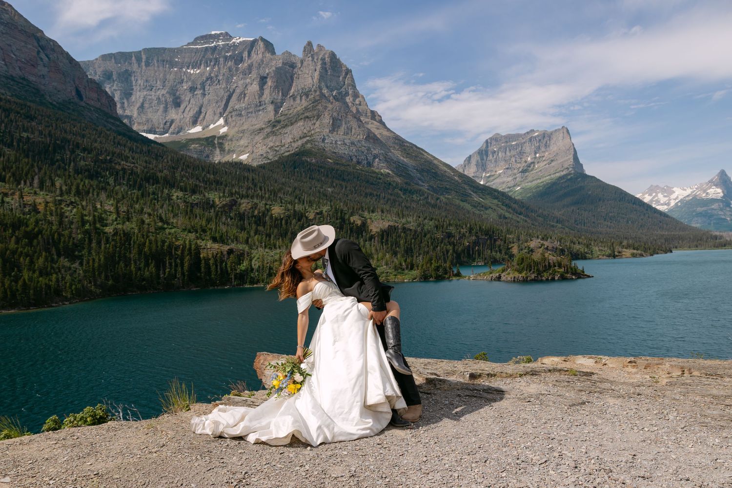 Bride and groom sit on rocky ledge overlooking turquoise mountain lake with dramatic peaks in Glacier National Park.