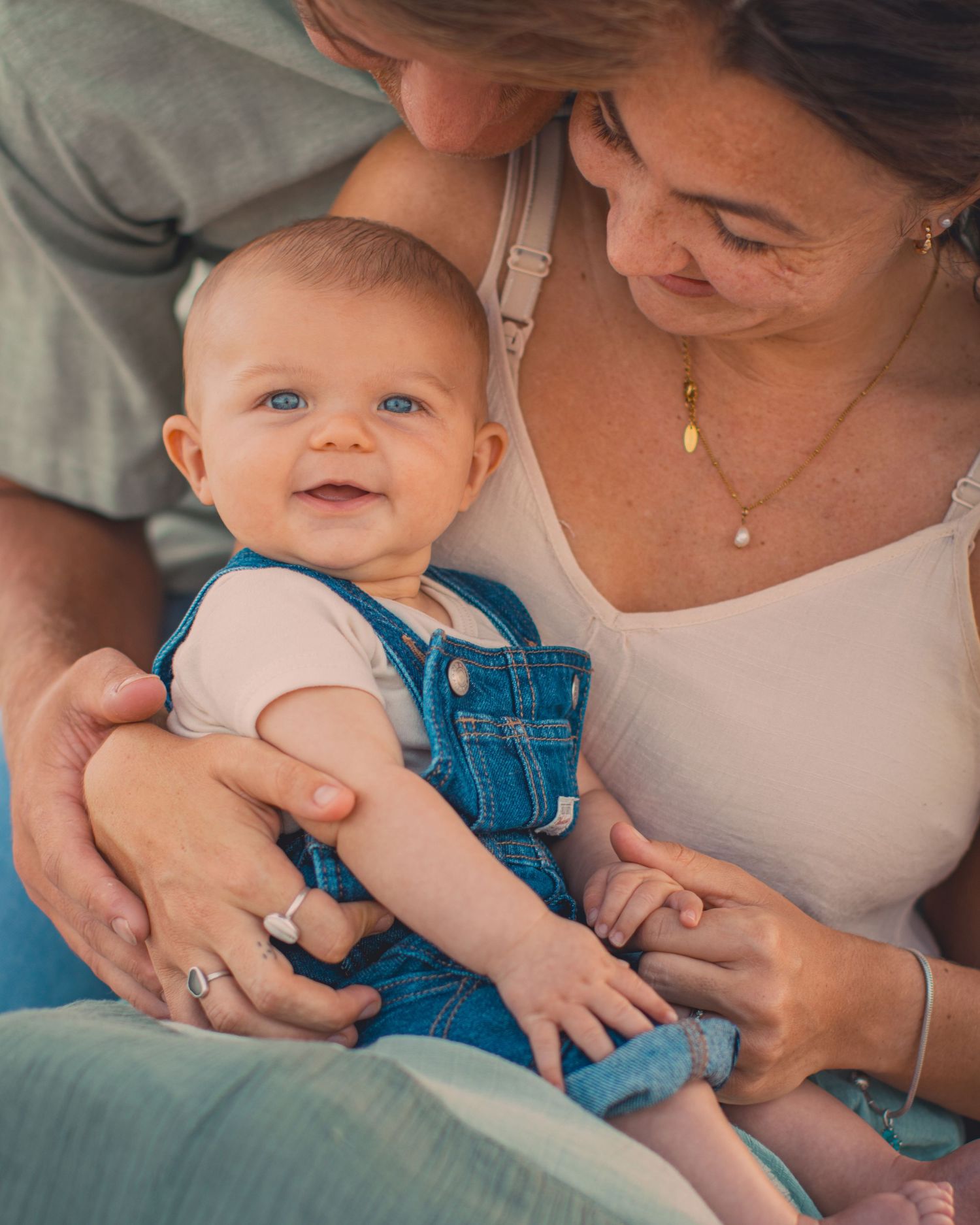Happy blue-eyed baby in denim overalls smiling while being held by parents in a warm family portrait.