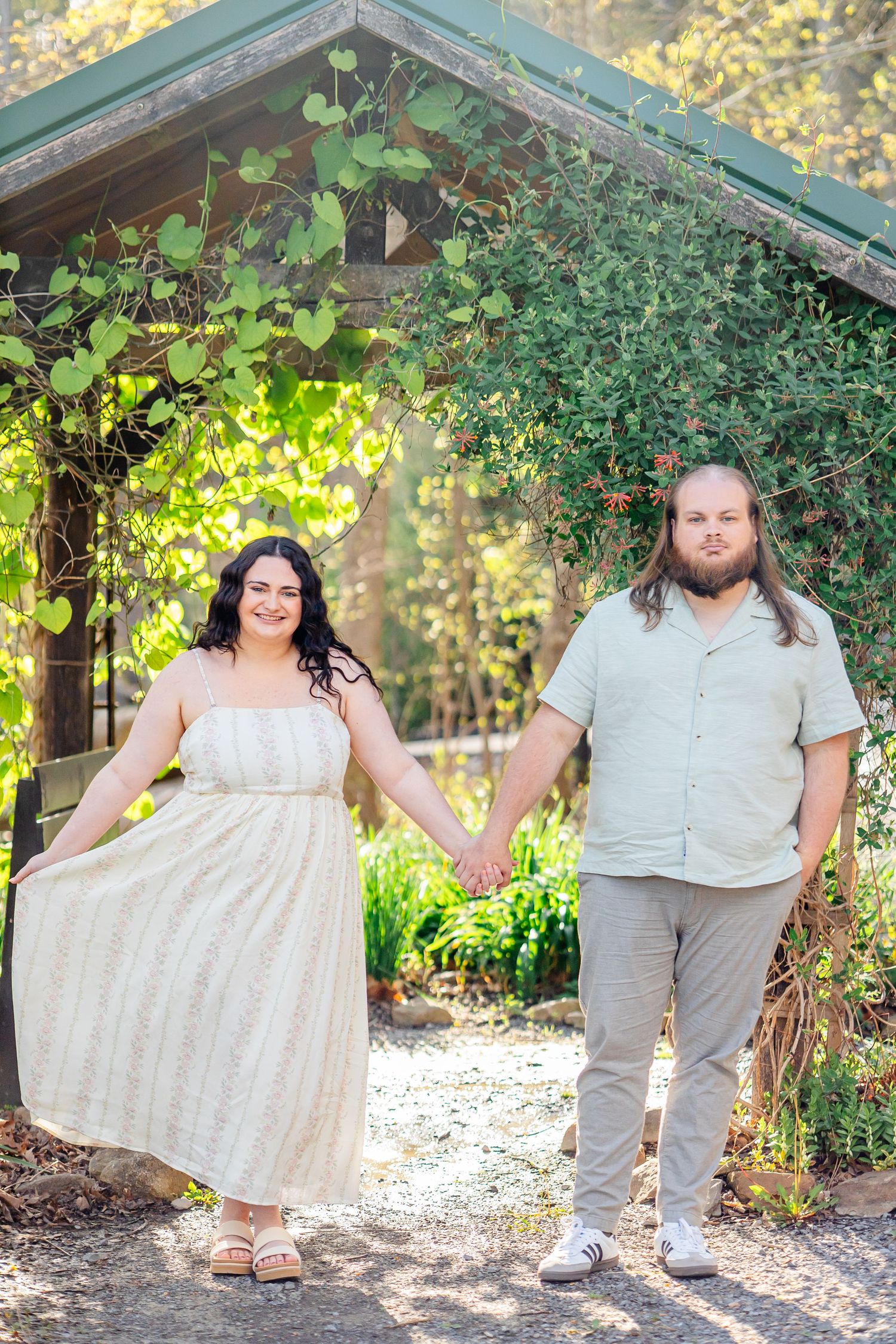 A couple holds hands smiling in a lush garden setting, woman in cream dress, man in casual attire, blue shirt and grey pants. morgantown wv