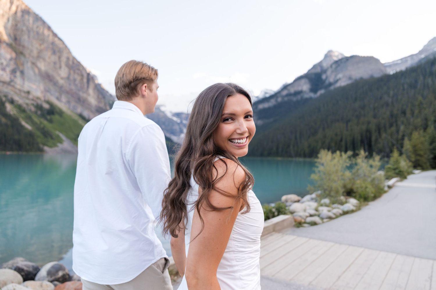 A smiling woman in a white dress poses with her partner by turquoise Lake Louise with mountain scenery backdrop.