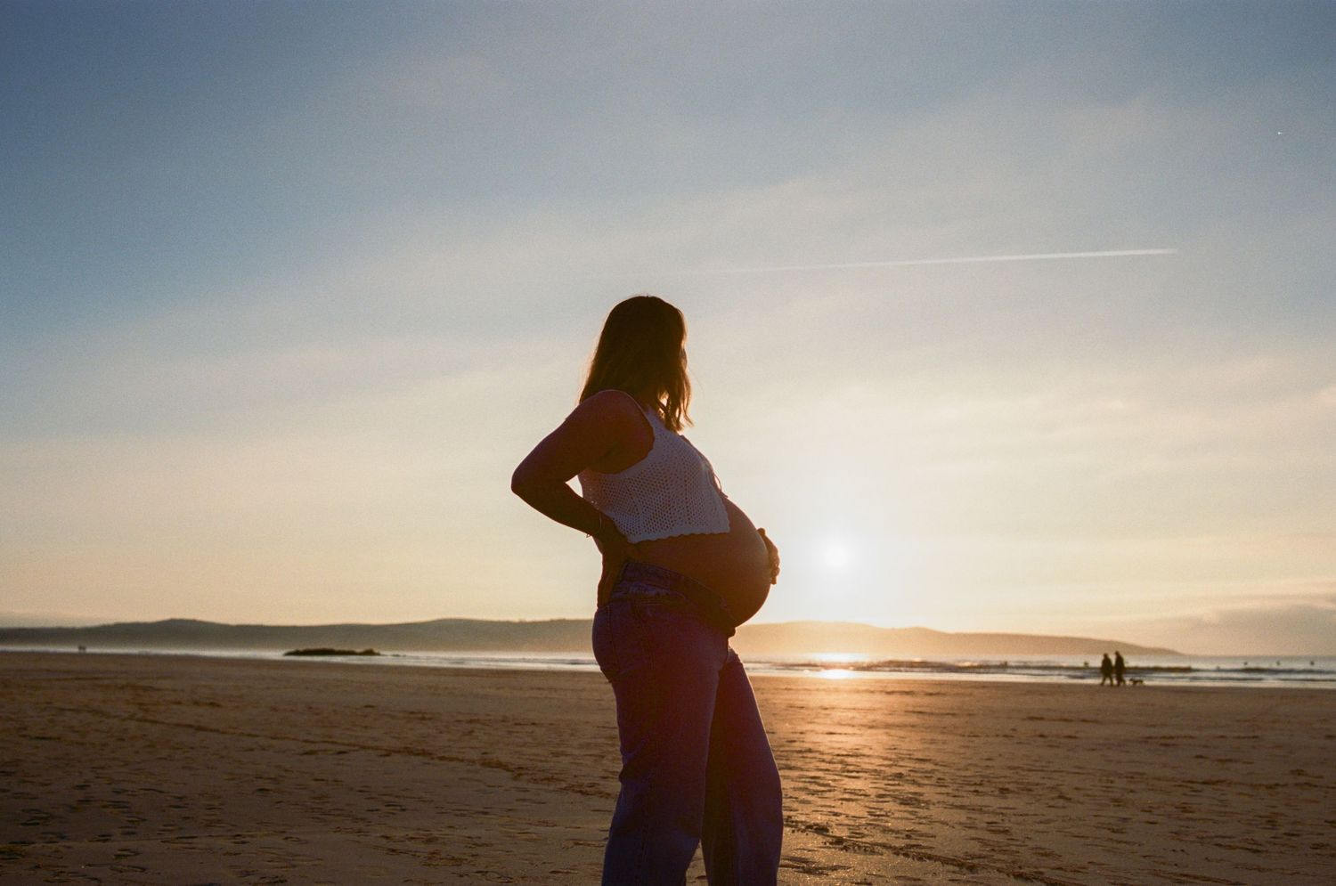 Pregnant woman silhouetted against golden sunset on sandy beach, cradling baby bump while gazing at ocean horizon.