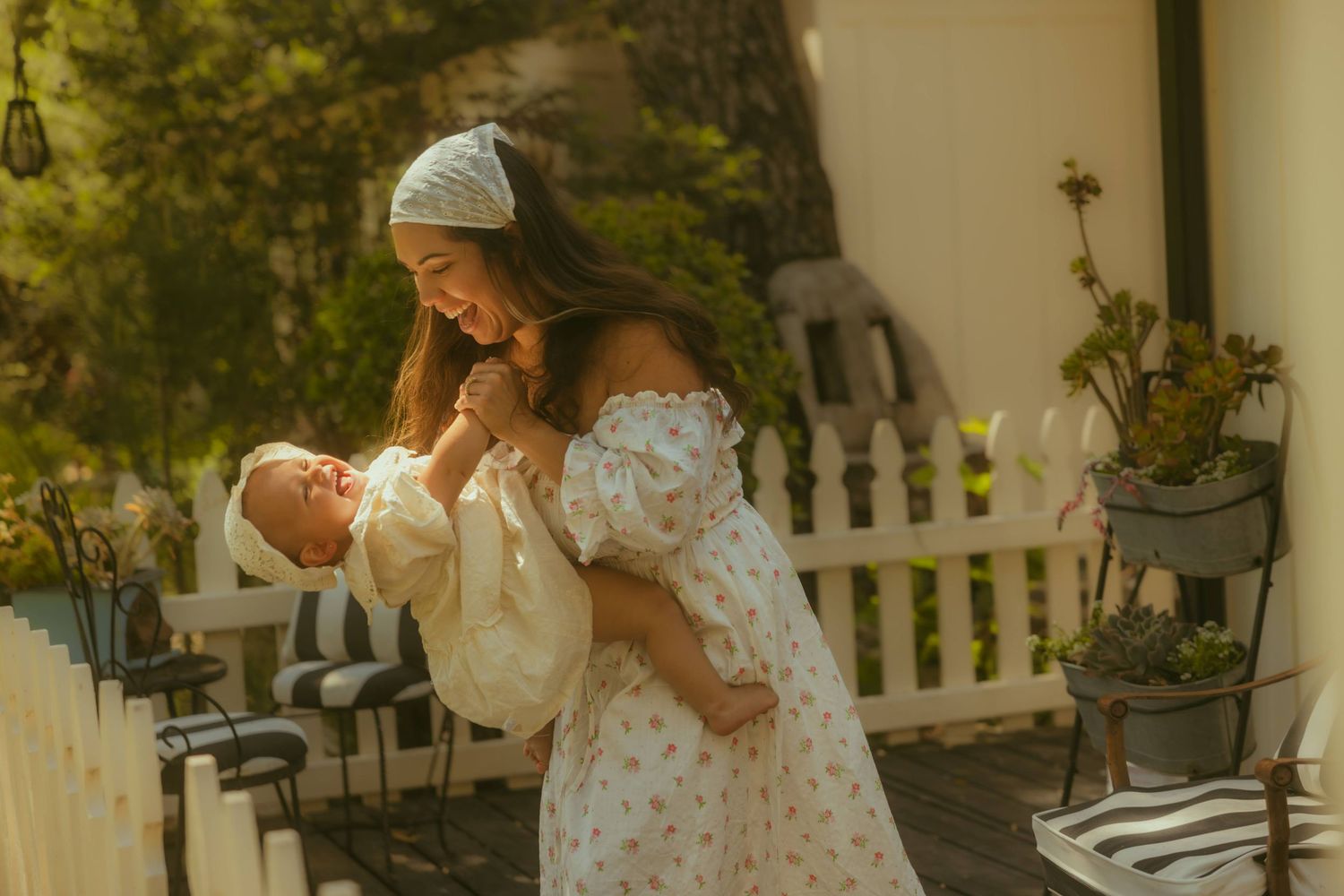 A young mother in a floral dress lovingly holds her newborn baby on a sunlit wooden porch photographer by Yoyo Leung