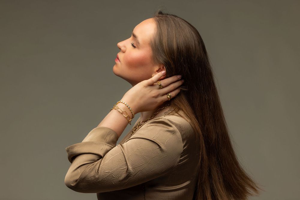 Woman in beige jacket with gold bracelet poses with eyes closed against gray background.