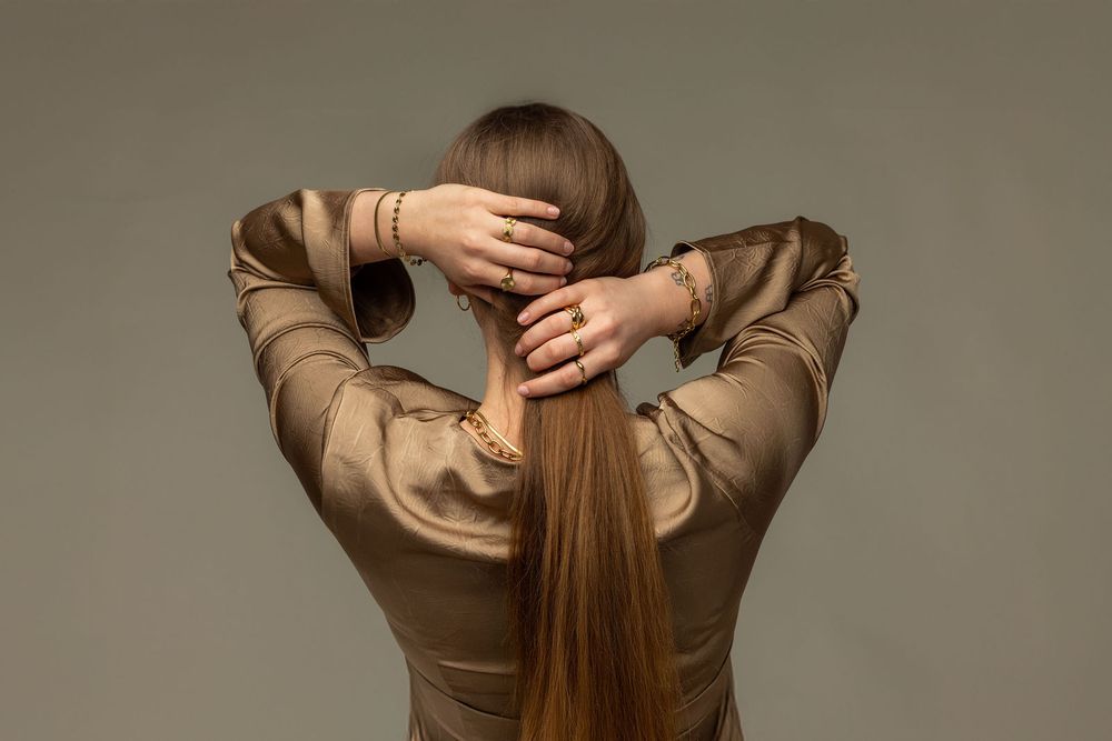 Woman in gold satin blouse holding her long straight brown hair back with both hands, wearing gold bracelets.