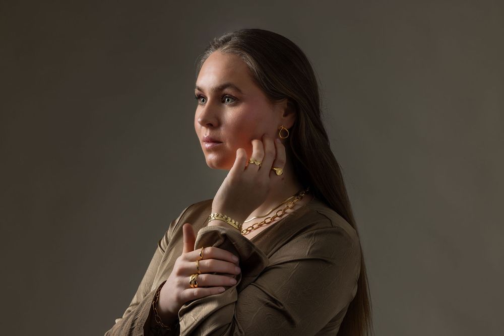 Woman in olive jacket with gold jewelry poses thoughtfully against gray background.