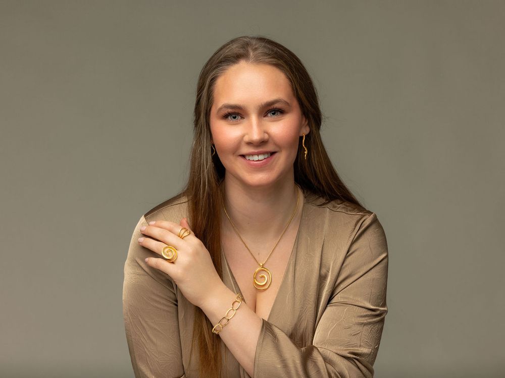 Smiling young woman with long hair wearing gold jewelry and a beige blazer against a gray background.