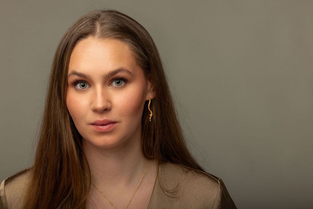 Young woman with long brown hair wearing gold earrings and necklace, posing against a gray background.