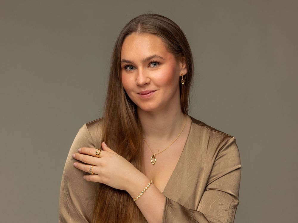 Young woman with long brown hair wearing gold jewelry and a beige top, posing gracefully against a gray background.
