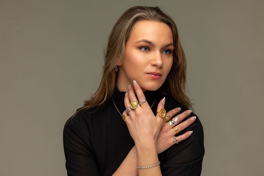 Woman modeling gold rings and jewelry in black turtleneck against gray background.