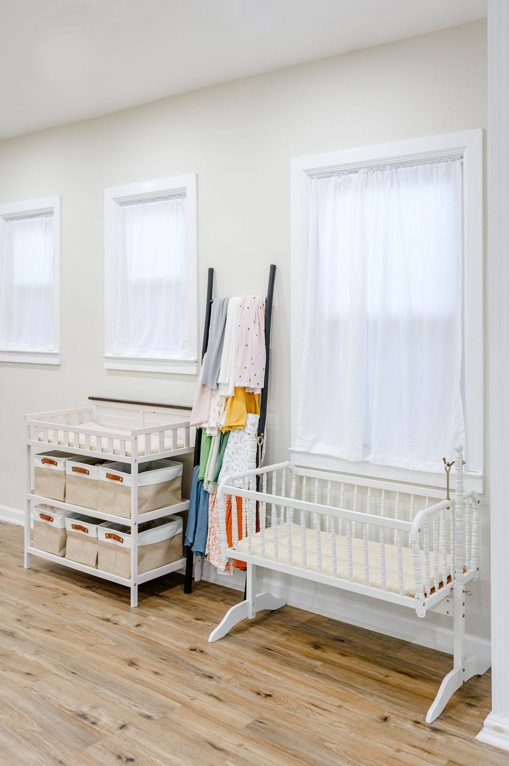 White nursery room with a changing table, storage baskets, a cradle, and a clothing rack with colorful baby clothes.