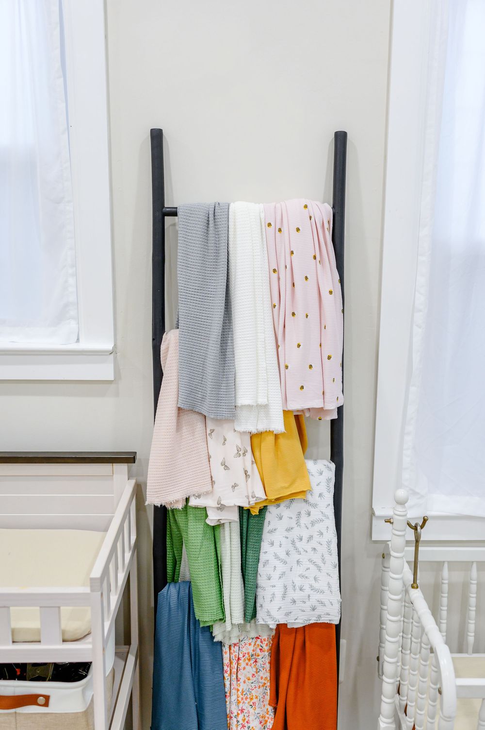 Colorful baby blankets and swaddles displayed on a black ladder rack between two white cribs in a nursery.