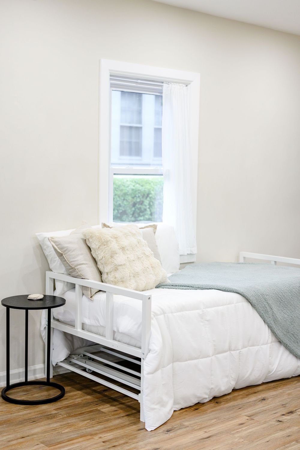 Minimalist white bed frame with soft pillows and sage green throw blanket beside a black side table near a sunlit window.