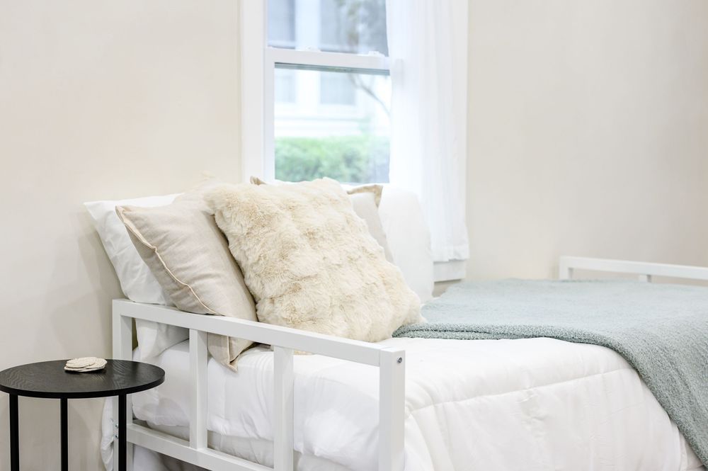 White bed with decorative pillows near a sunlit window, featuring a mint green blanket and minimalist black side table.