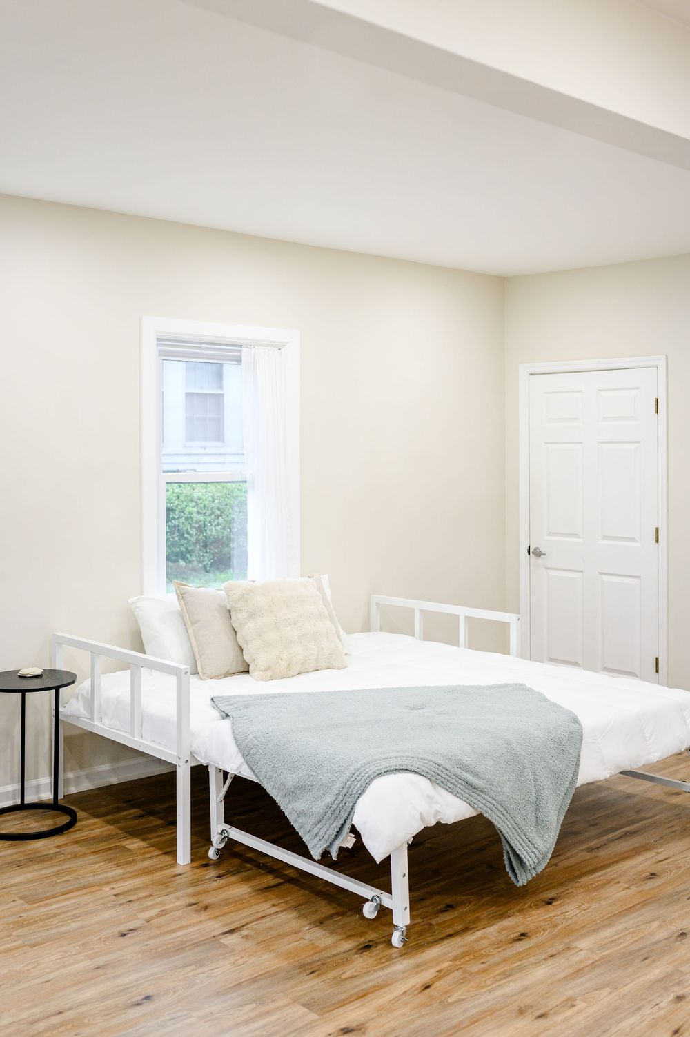 Minimalist bedroom with white metal daybed, sage green throw blanket, hardwood floors, and neutral walls with natural window light.
