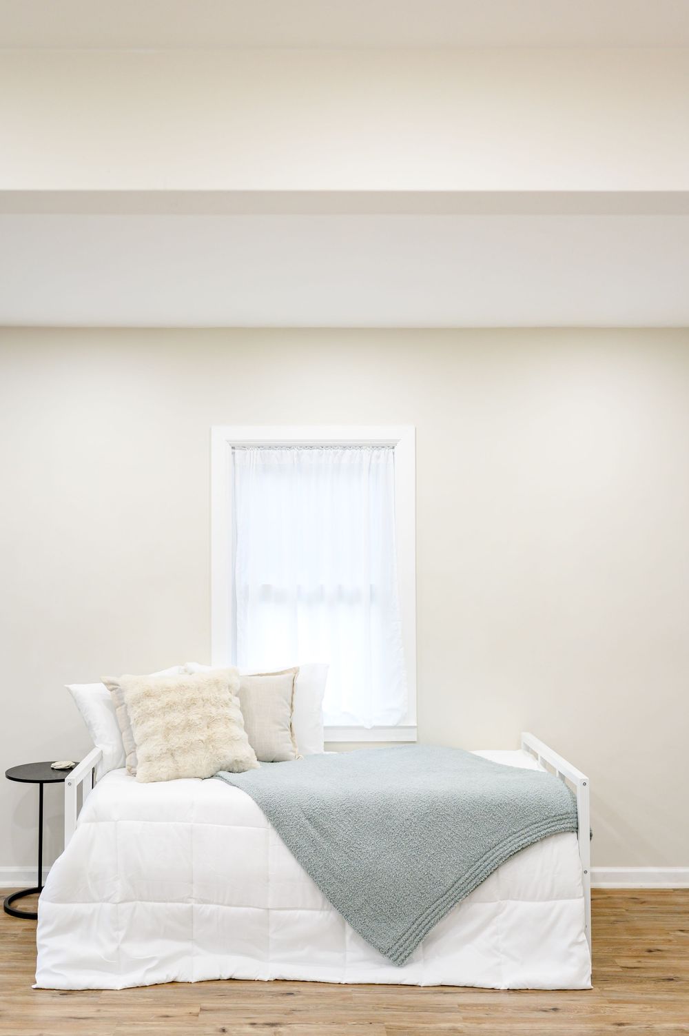 Minimalist white bedroom with a daybed, soft pillows, light blue throw blanket, and a bright window with sheer curtains.