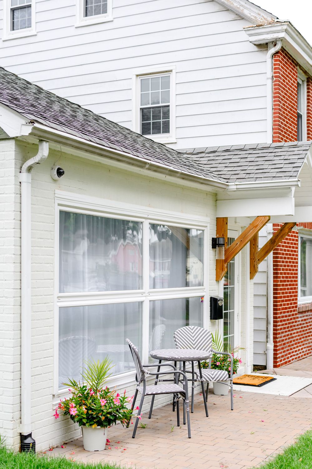 Cozy home patio with bistro table, chairs, potted flowers, and large sliding glass doors on a brick and white siding house.