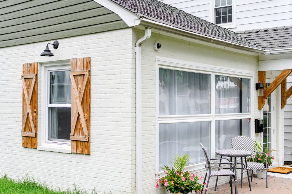 White brick cottage exterior with rustic wooden shutters, large window, and charming patio seating area surrounded by lush greenery.