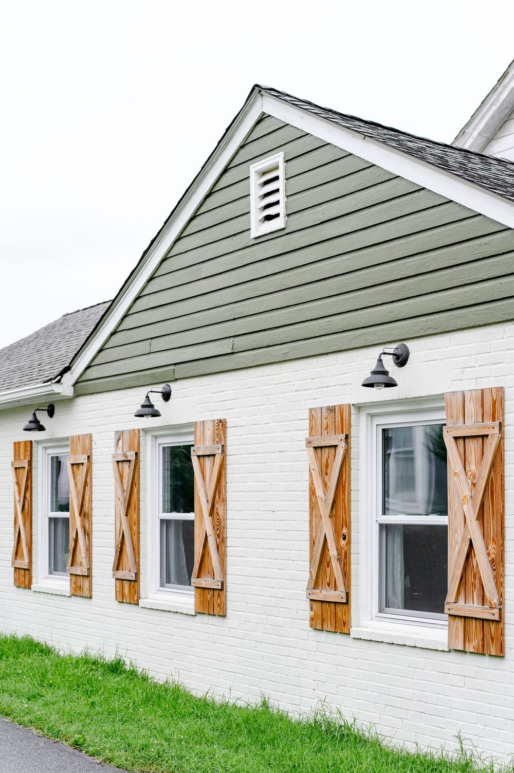 White brick farmhouse with rustic wooden shutters, sage green siding, gable roof, and black wall sconce lights.