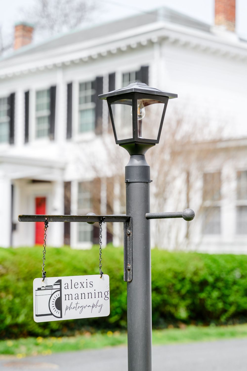 A lamp post with an Alexis Manning Photography sign hanging from it, in front of a white colonial house.