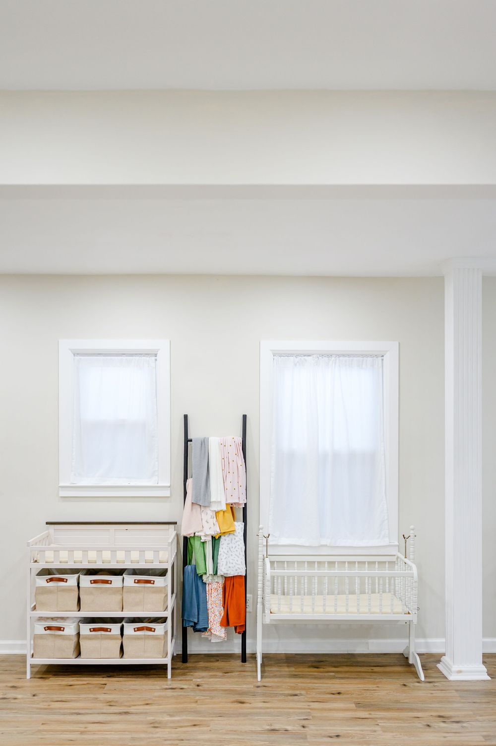 White nursery with changing table, colorful clothing rack, and vintage crib between two bright windows on hardwood floors.