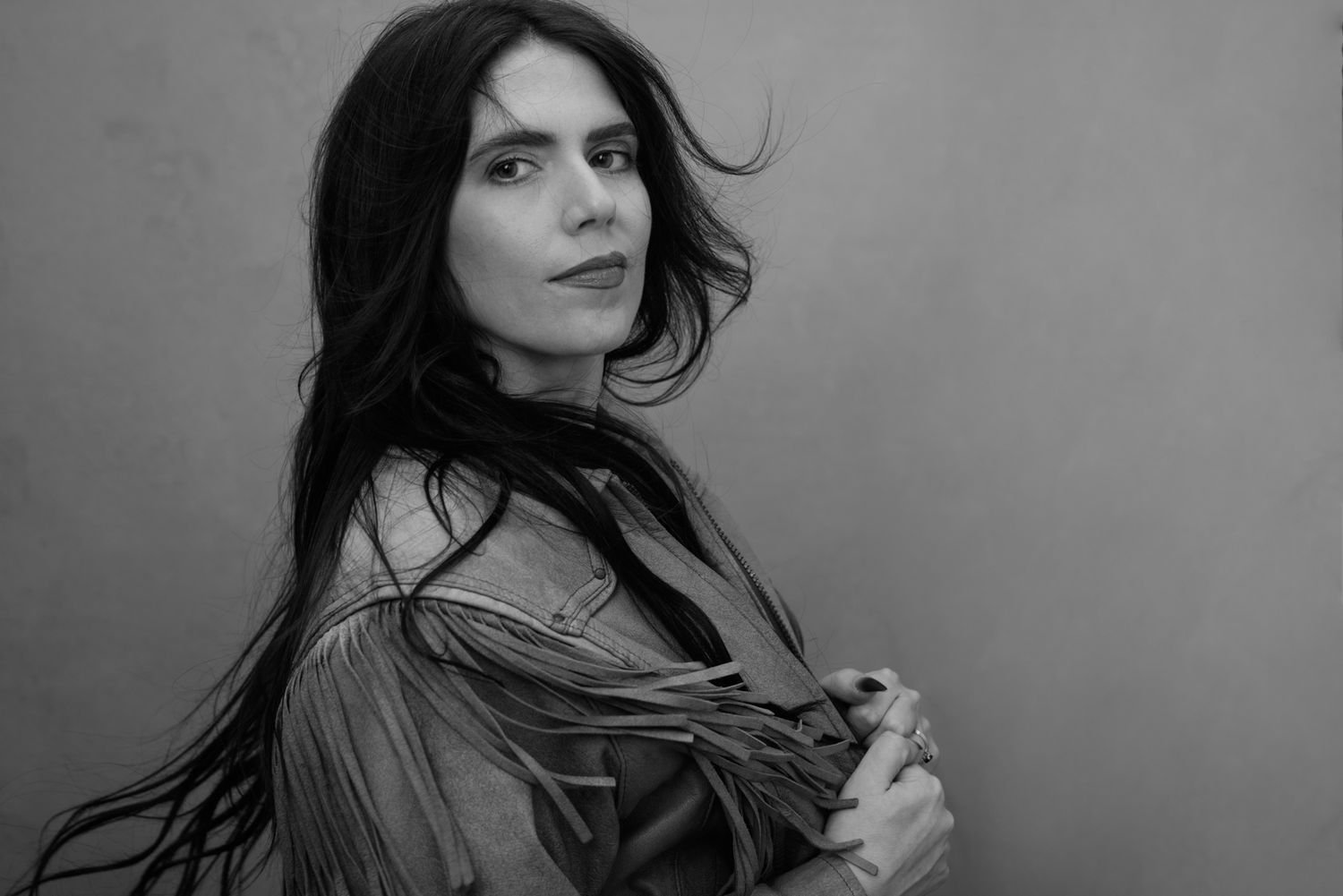 Black and white portrait of a woman with long dark hair, tattoos, posing with hand behind head.