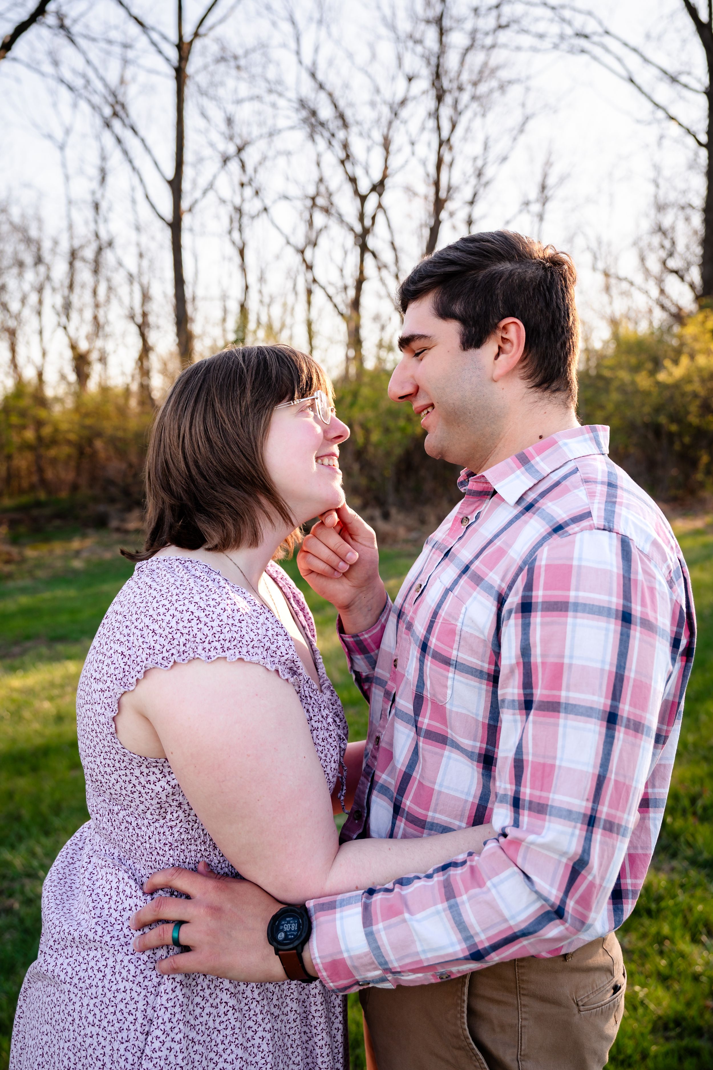 A couple shares a tender moment outdoors, the man gently touching the woman's chin as they smile at each other.