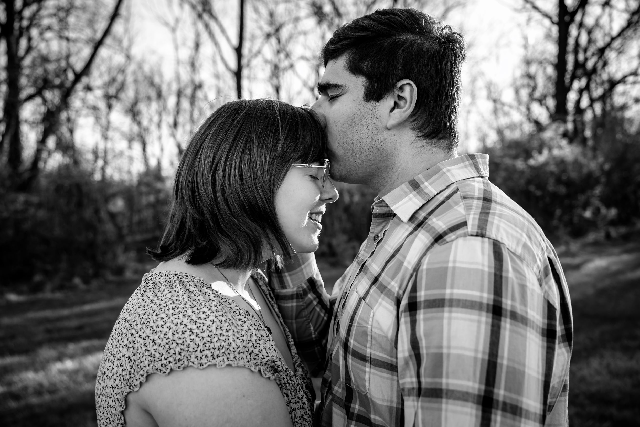 Black and white photo of a couple sharing a tender moment outdoors among bare trees.