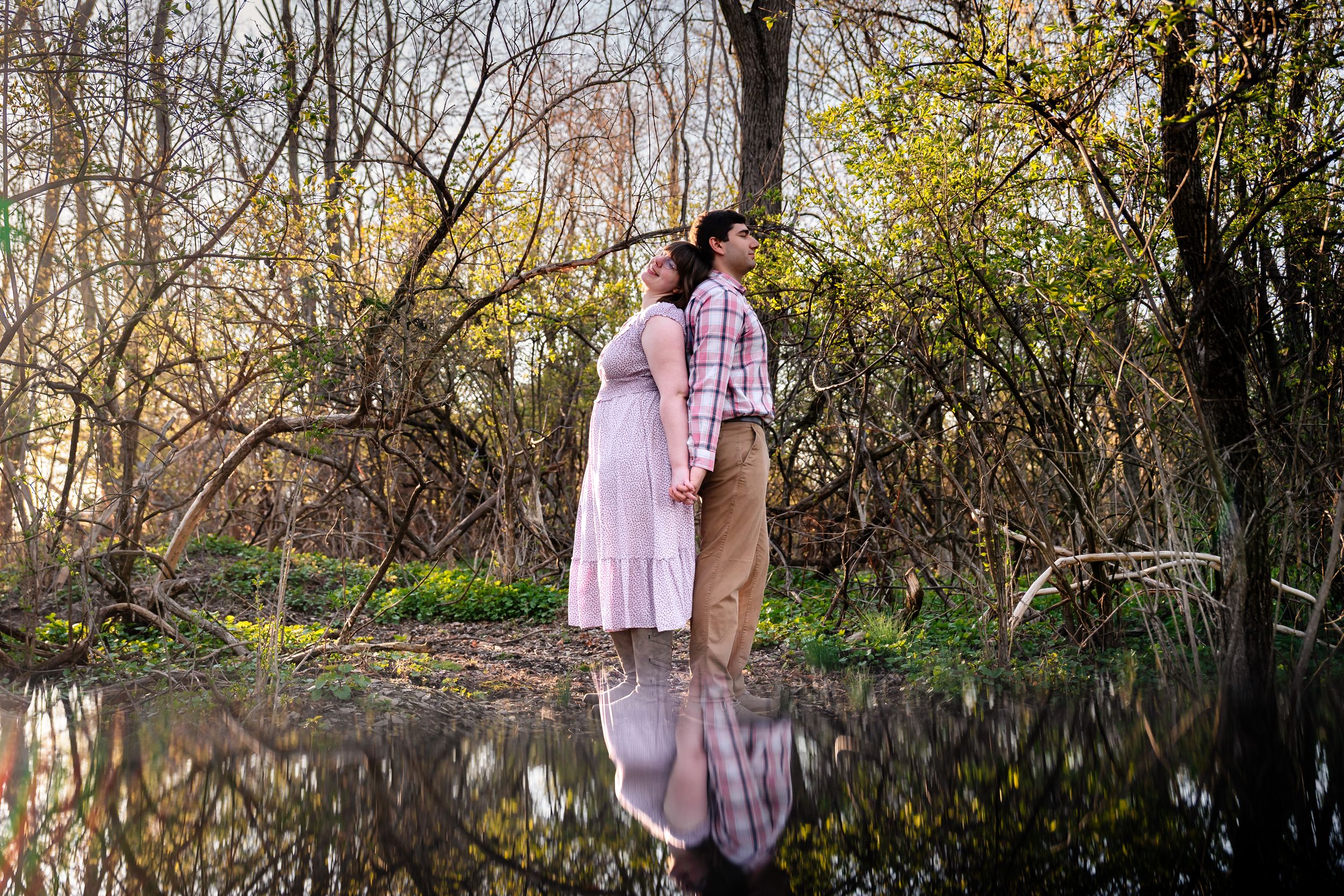 Couple stands back-to-back near a reflective pond surrounded by trees in early spring.