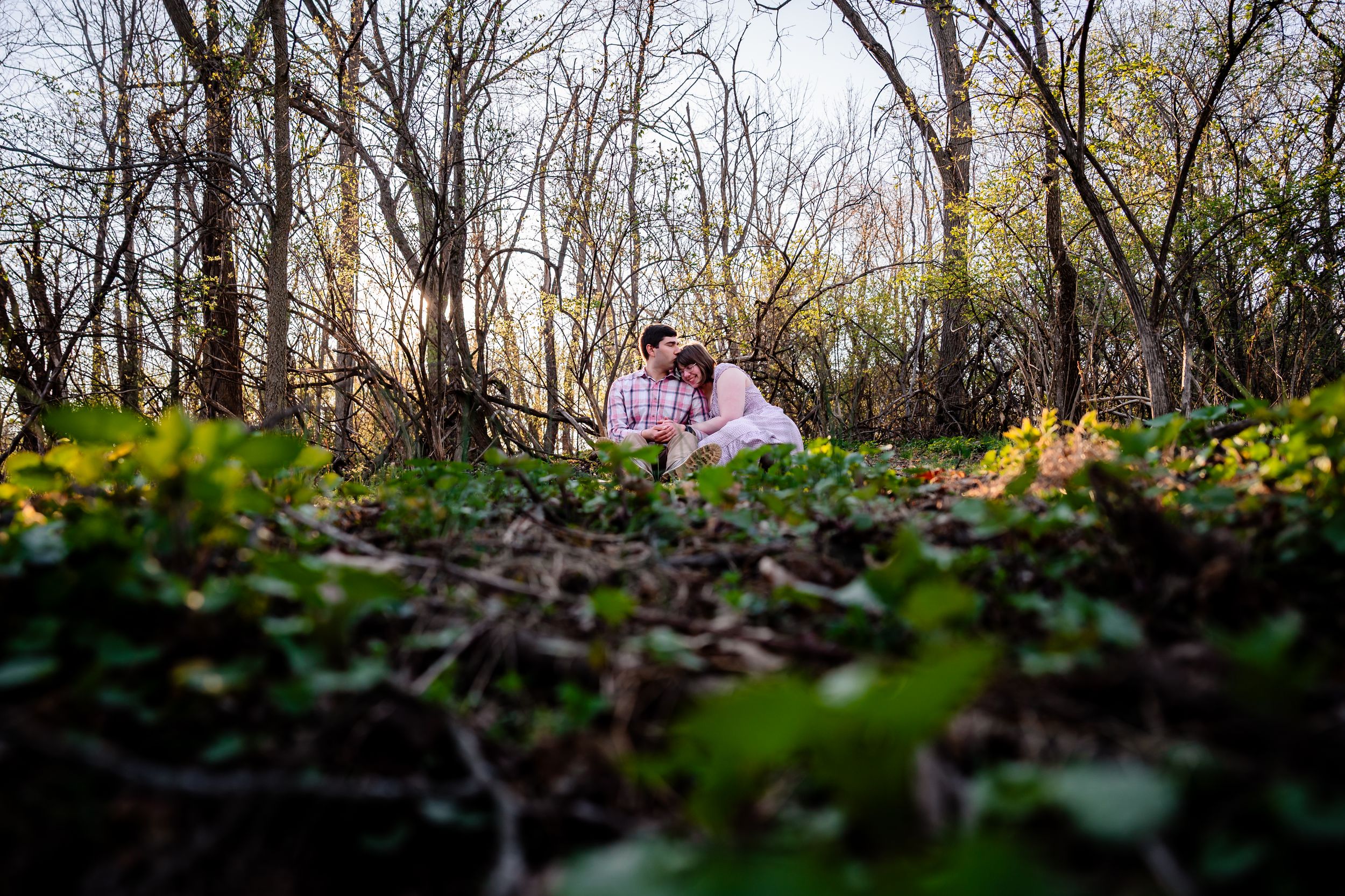 Couple sitting together in a lush forest clearing surrounded by green plants and bare trees in spring.