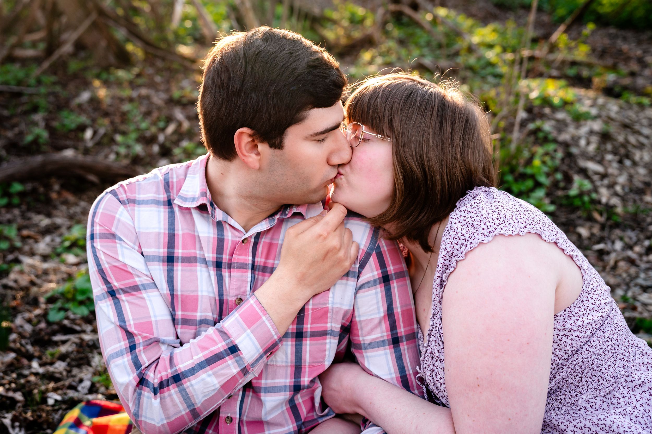 A couple shares a romantic kiss outdoors during a sunny engagement photo session in a natural wooded setting.
