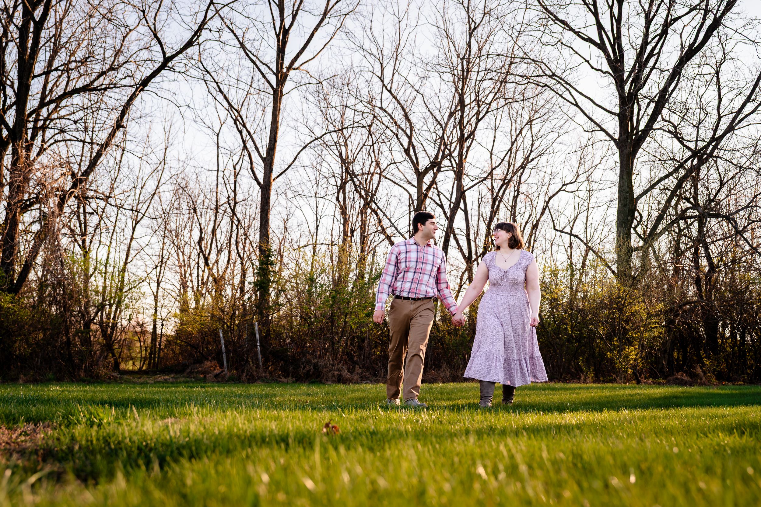 Couple holding hands walking on green grass in a park with bare trees at golden hour.