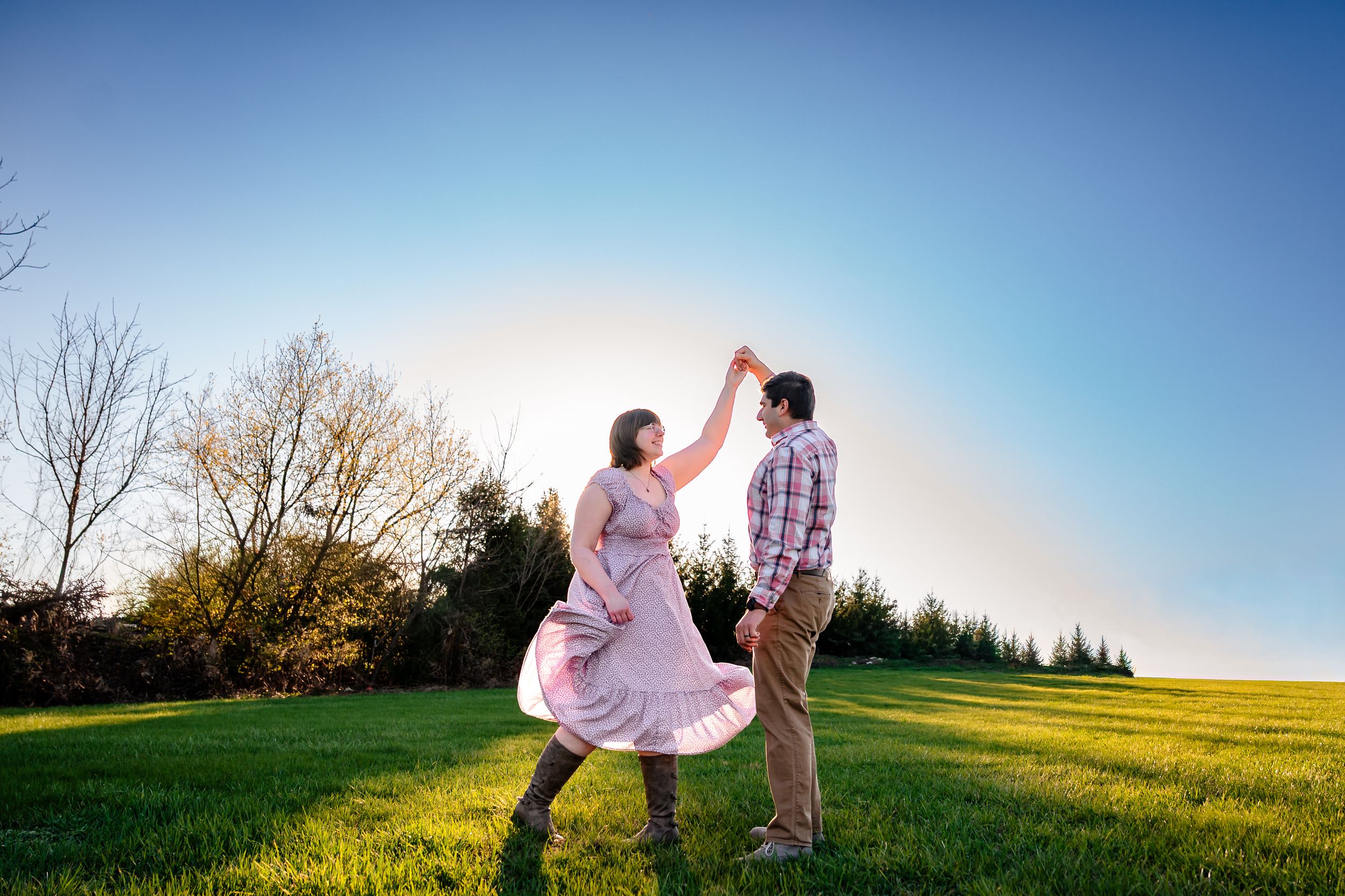 Couple dancing together in a lush green field at sunset, woman in pink dress twirling under man's raised hand.