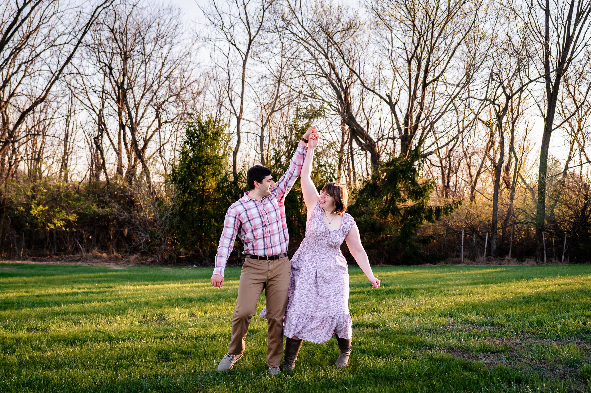 Happy couple dancing together in a green field surrounded by bare trees at golden hour.