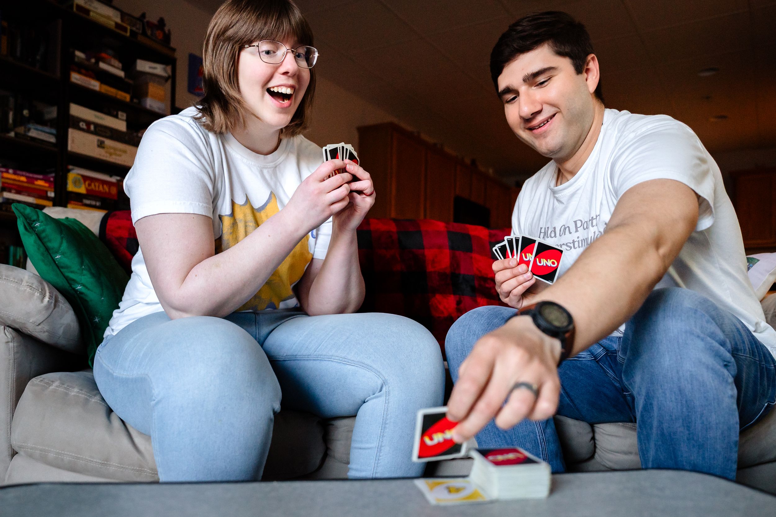 Two people laughing and playing Uno card game together on a couch indoors.