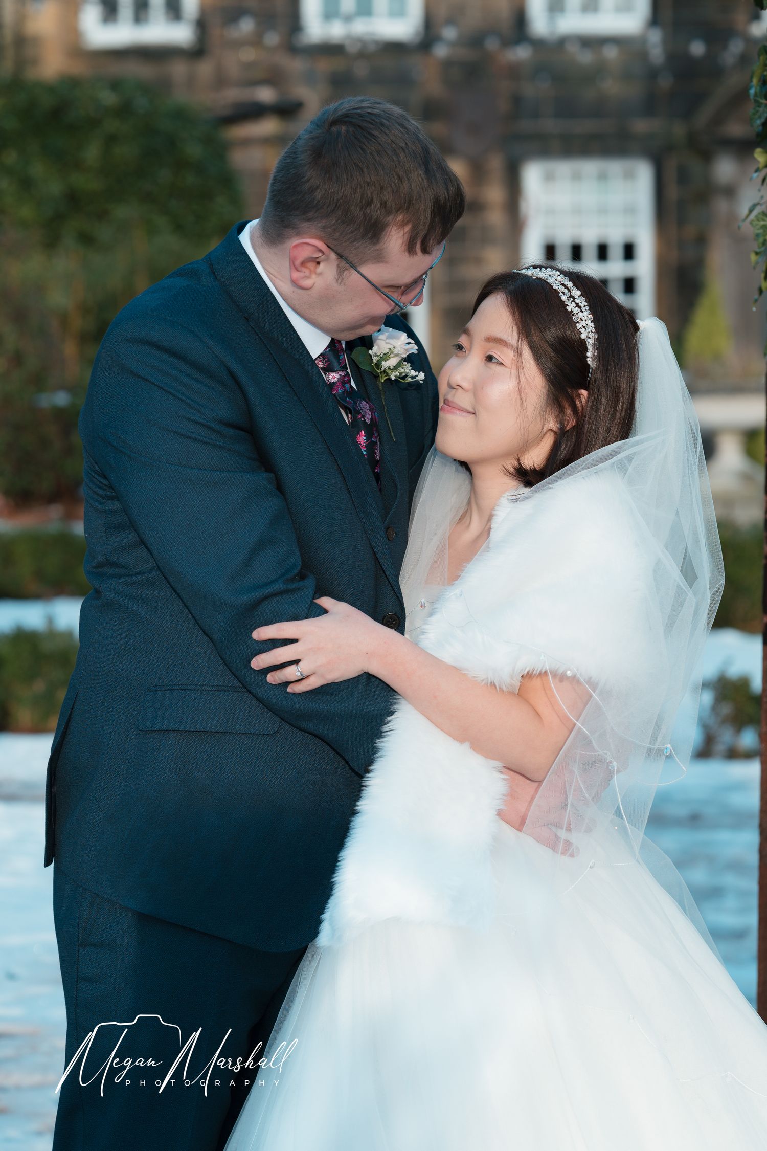 Bride and groom share a romantic kiss outside a stone building, bride wearing white gown with veil and fur wrap.