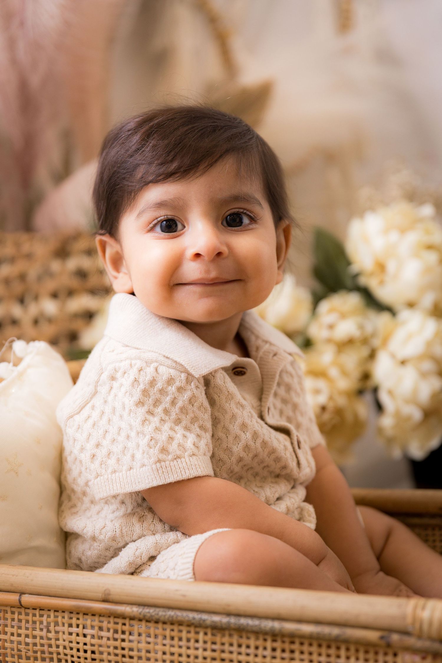 Smiling baby in a cream knit outfit sitting in a wicker basket with white flowers in the background.