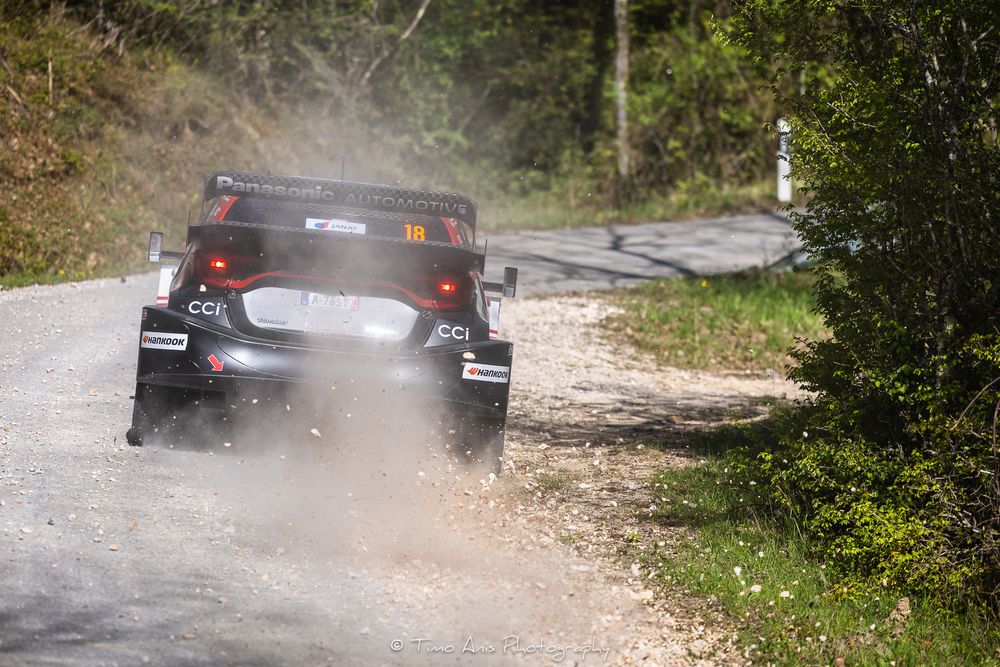 Rally car with CCI livery speeding on a dusty gravel road surrounded by trees during a rally stage.