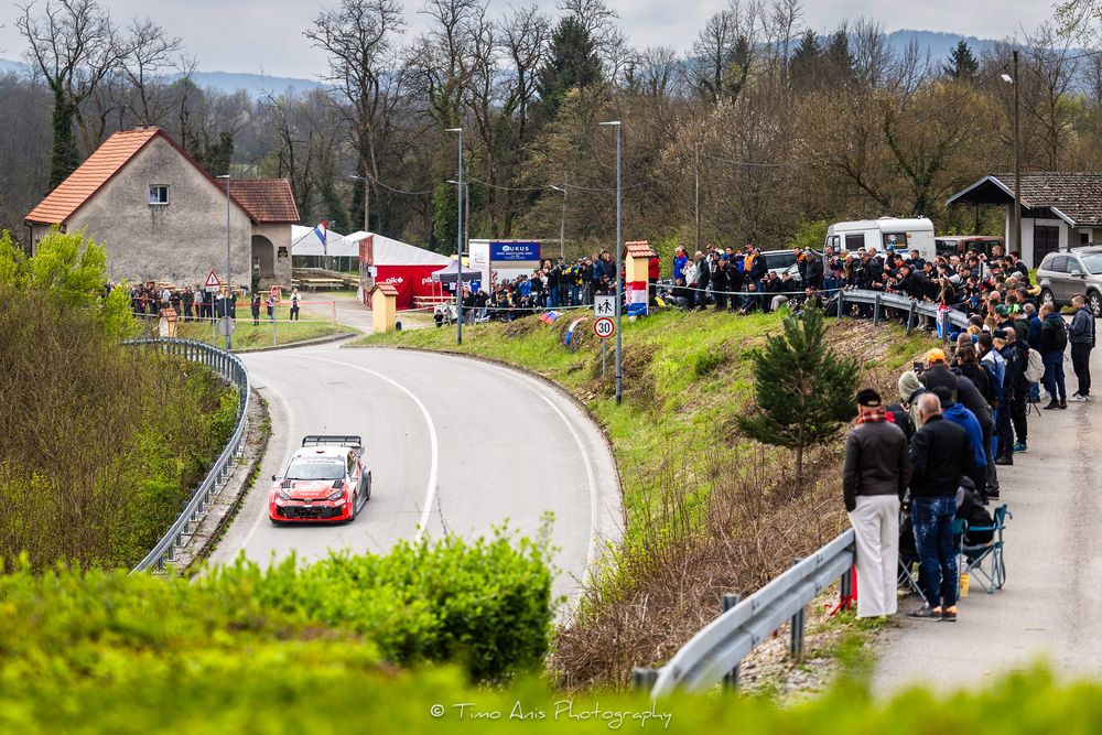 Spectators lining a curved rally stage road watching a white race car compete in a rally event.