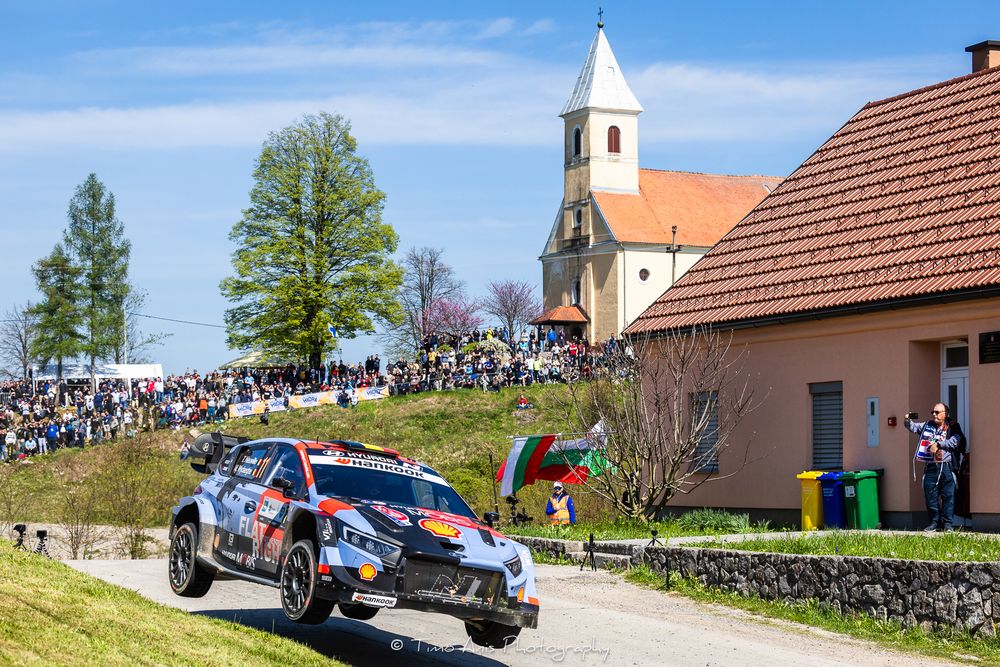 A rally car races past a church and crowd of spectators on a gravel road during a WRC rally event in sunny conditions.