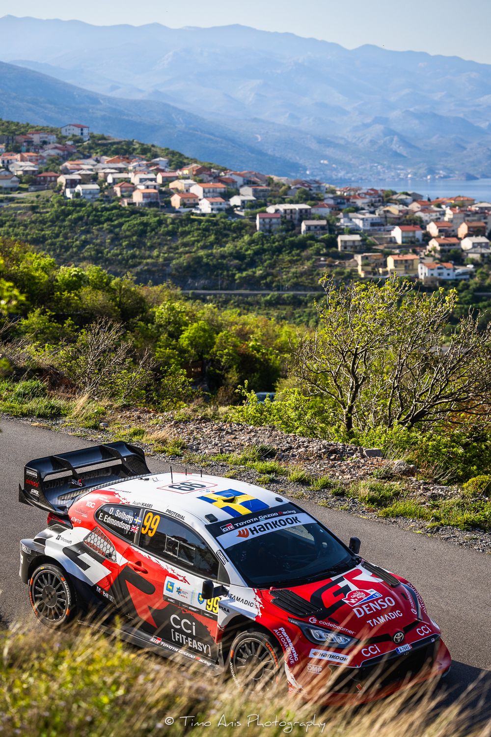 Toyota GR Yaris rally car number 99 racing on a mountain road with a scenic village and misty hills in the background.