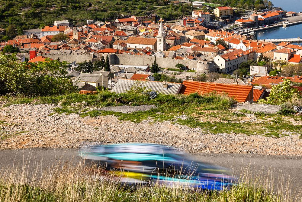 Colorful boats blur in motion on a hillside above a scenic Croatian coastal town with red-roofed buildings.