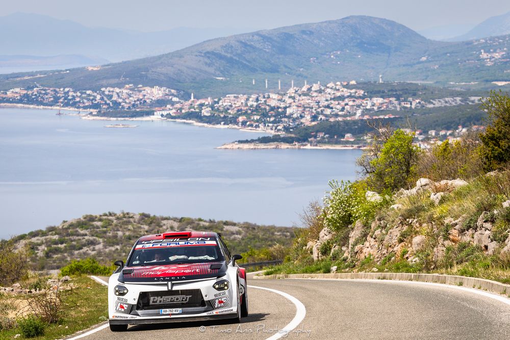 Citroen C3 WRC rally car racing on a coastal road with a scenic Mediterranean town and blue sea in the background.