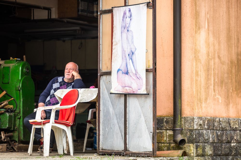 Man sitting on white chair outside a building beside a framed figure painting on a rustic wall.