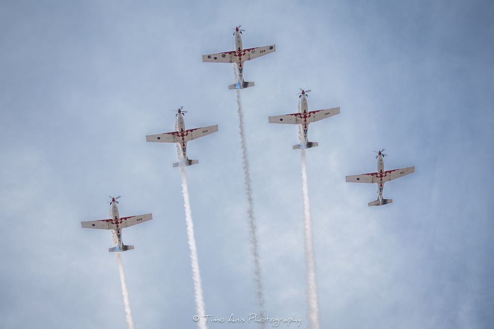 Five military aircraft fly in formation emitting white smoke trails against a cloudy blue sky at an airshow.